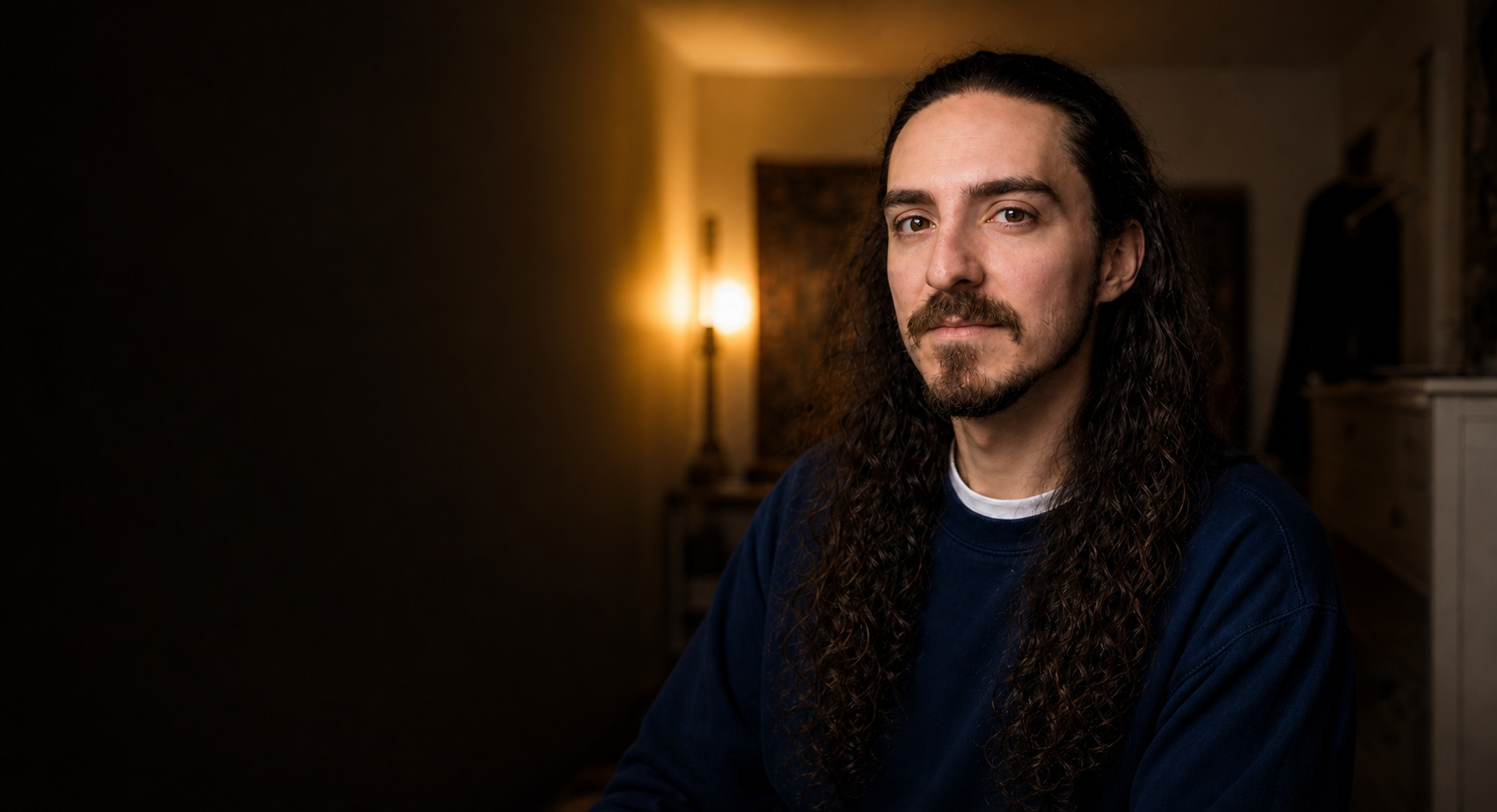 A man with long curly hair and a goatee, wearing a navy blue shirt, sitting indoors with soft lighting in the background.