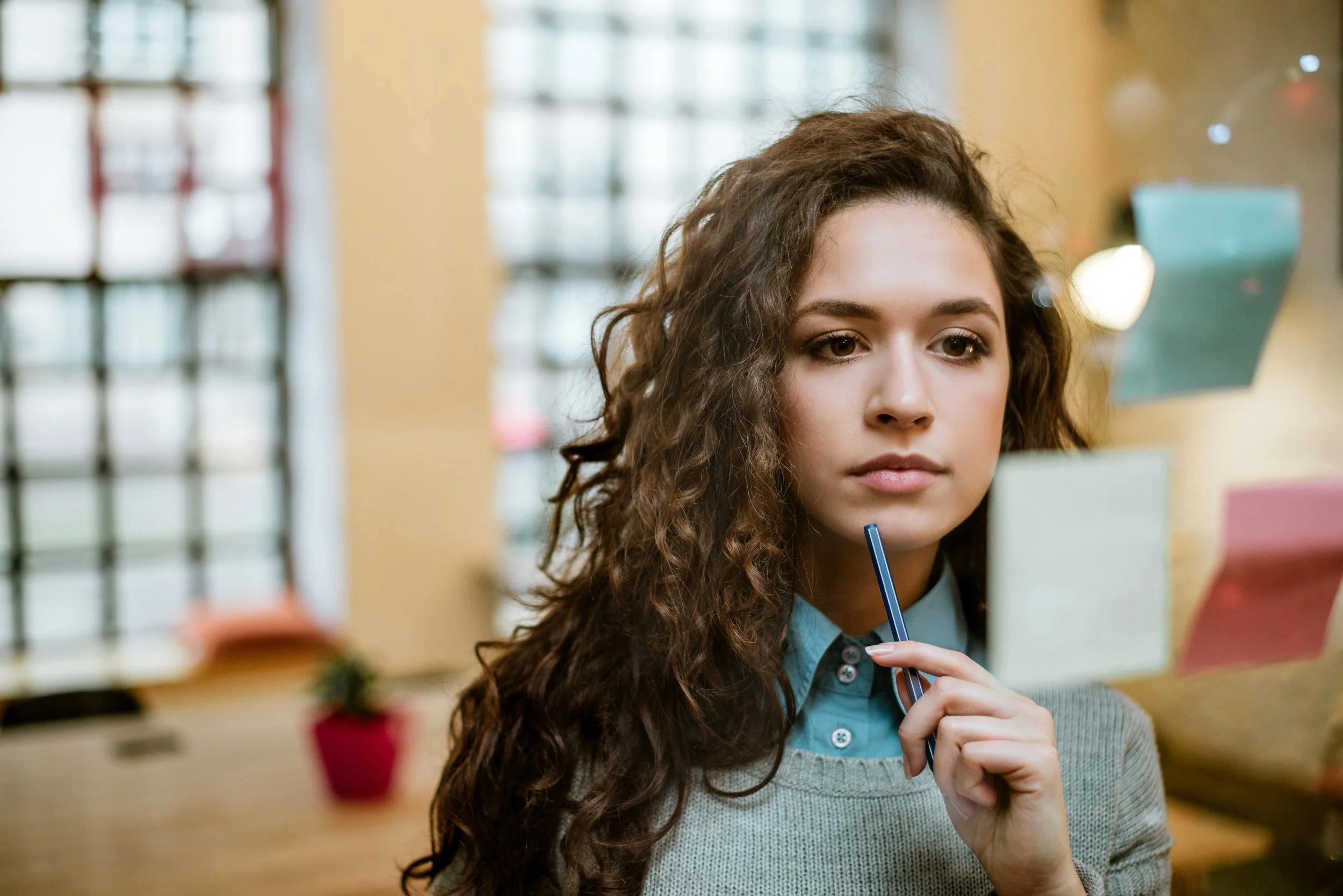 Young woman with curly brown hair in an office, holding a pen to her chin, looking thoughtfully at a computer screen.