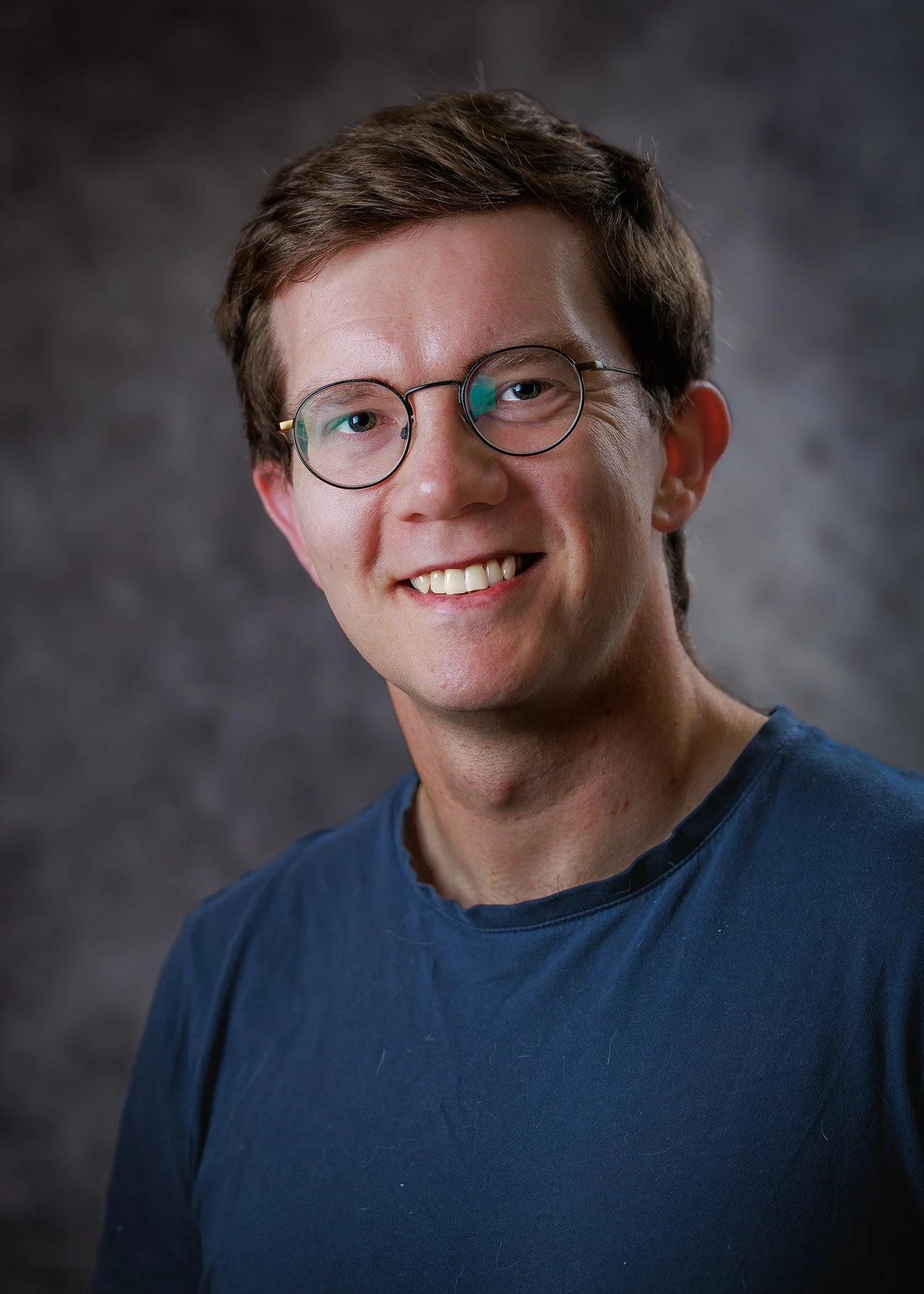 A young man with short brown hair, glasses, and a friendly smile wearing a blue t-shirt.