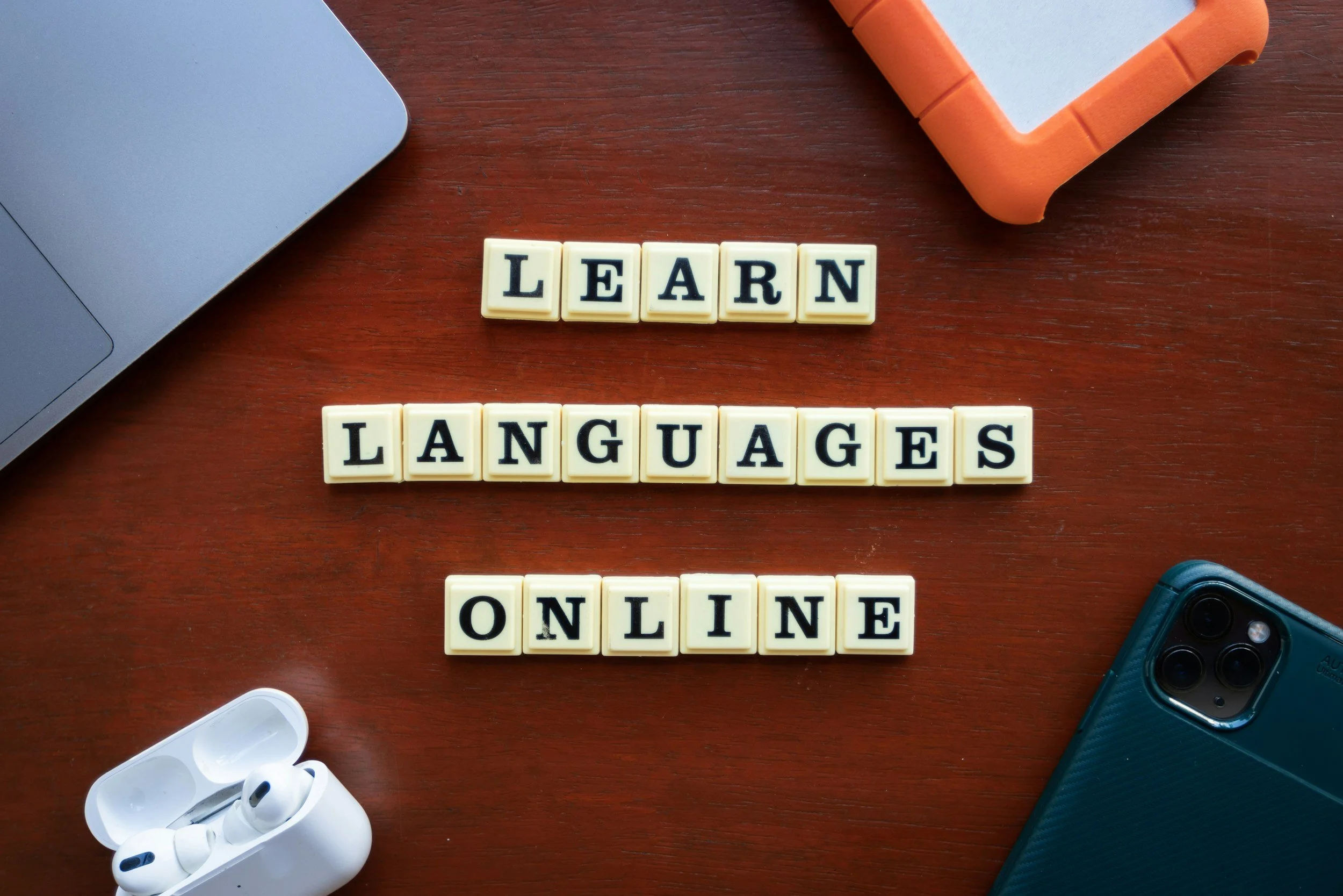 Scrabble tiles spelling 'LEARN LANGUAGES ONLINE' placed on a wooden desk next to a laptop, wireless earbuds, an orange frame, and a blue phone.