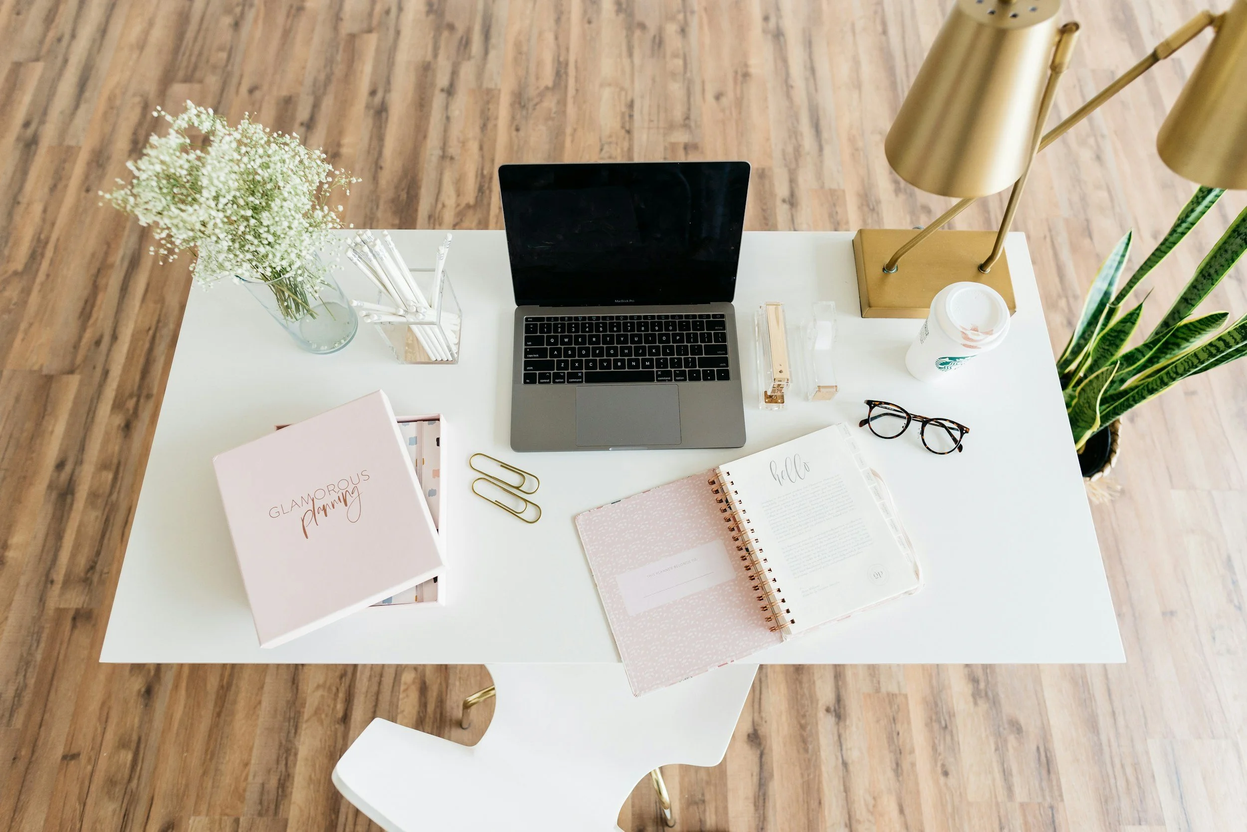 A white desk with a laptop, notebooks, a phone, a flower vase, a coffee cup, glasses, and office supplies, with a wooden floor in the background.