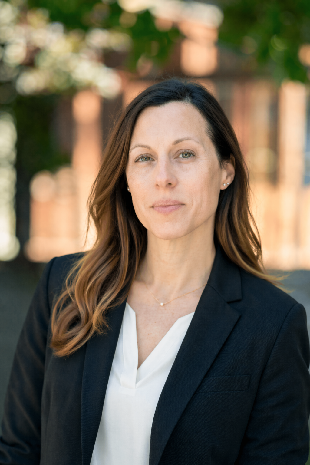 Professional woman with brown hair in loose waves, wearing a black blazer and white blouse, standing outdoors with blurred green trees and a brick building in the background.