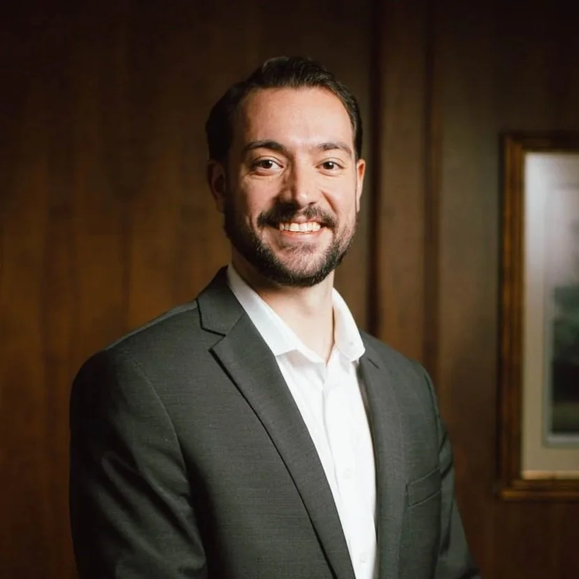 A man with a beard in a dark suit and white shirt smiling in a professional indoor setting with wood-paneled walls and a framed picture in the background.