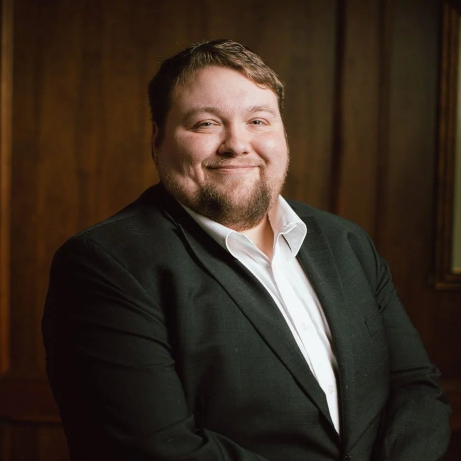 A man with short brown hair, a beard, and a smiling expression wearing a black suit and white shirt, standing in front of a wooden-paneled background.