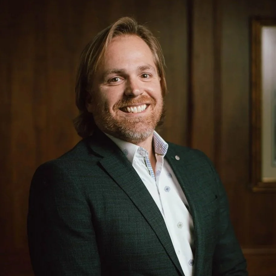 A smiling man with shoulder-length blonde hair, a beard, wearing a dark blazer and a patterned white shirt, standing in front of a wooden wall.
