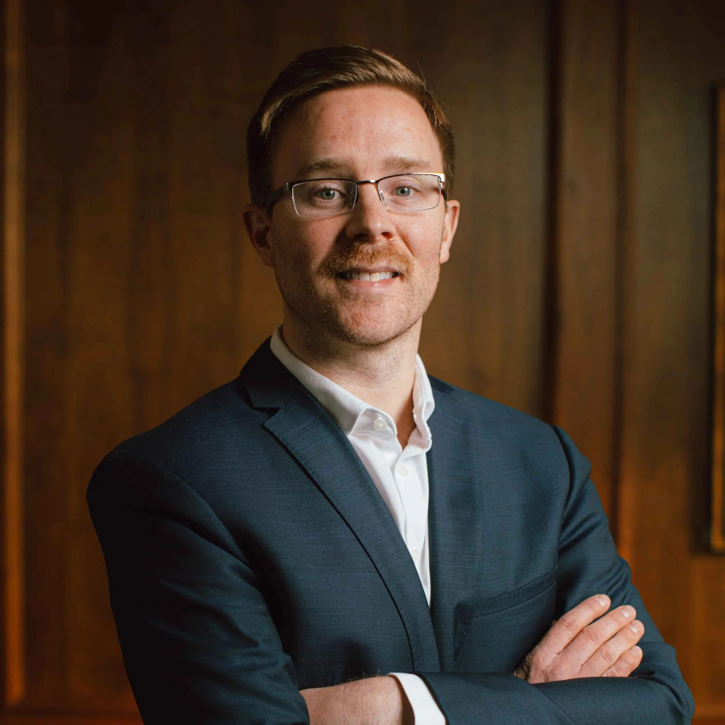 Close-up of a smiling man wearing glasses, a dark blue suit, and a white shirt, standing with arms crossed in front of a wooden background.