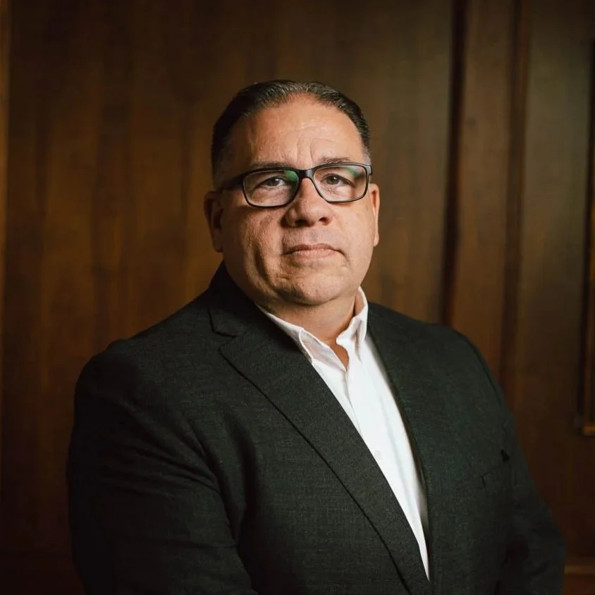 A middle-aged man with short, dark hair and glasses, dressed in a white shirt and dark blazer, standing against a wooden paneled wall.