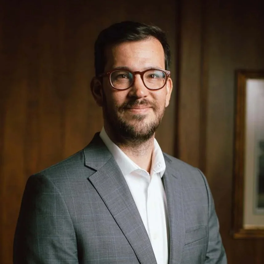 Portrait of a man wearing glasses and a gray suit with a white shirt, standing in front of a wooden wall.