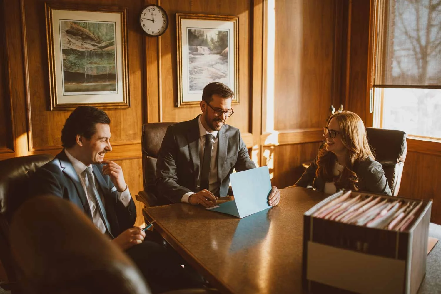 Three business professionals, two men and one woman, sitting at a wooden conference table in a wood-paneled office, smiling and engaging in a discussion. The man in the middle is reading a document, the woman is laughing, and the other man is holding a smartphone. There are framed landscape pictures on the wooden wall behind them, and a window with blinds on the right side, letting in natural light.