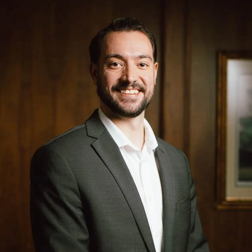 A man with dark hair and a beard smiling, wearing a gray suit and white shirt, standing in front of a wooden wall and framed picture.