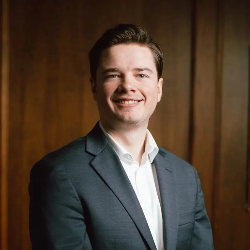 A young man in a gray suit and white shirt smiling in front of a wooden wall.