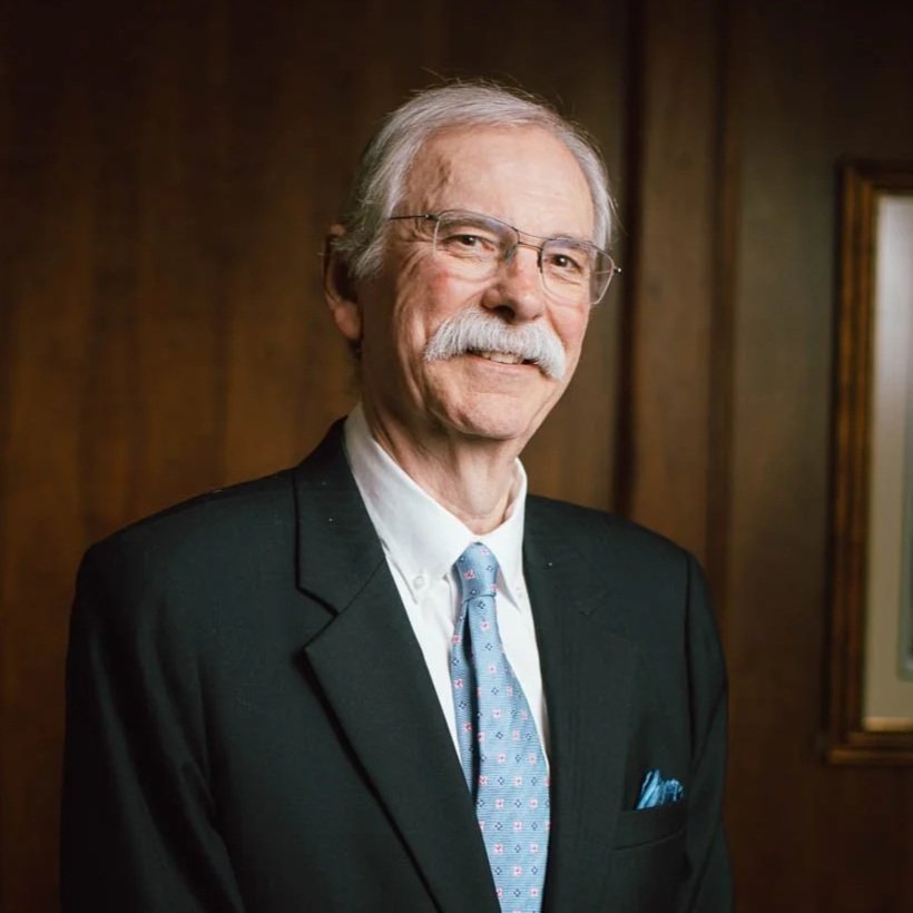 An elderly man with gray hair, glasses, and a mustache, wearing a black suit, white shirt, and blue patterned tie, standing indoors with a wooden wall background and framed art.