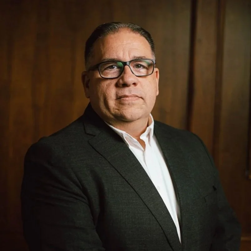 Portrait of a man wearing glasses in a dark suit with a white shirt, standing in front of a wooden background.