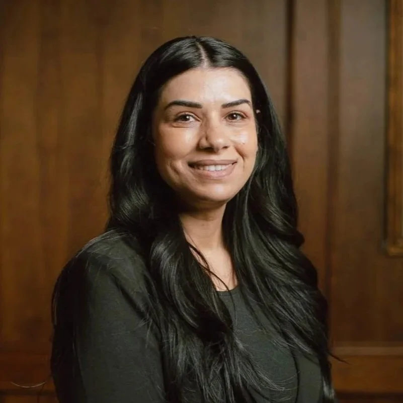 A woman with long black hair smiling at the camera, wearing a black top, against a wooden background.