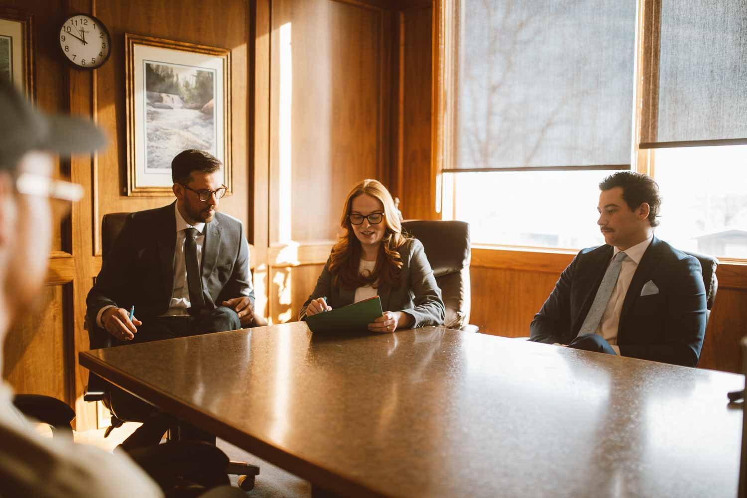Group of three professionals in business suits having a meeting in a wood-paneled conference room with a large window and artwork on the wall.