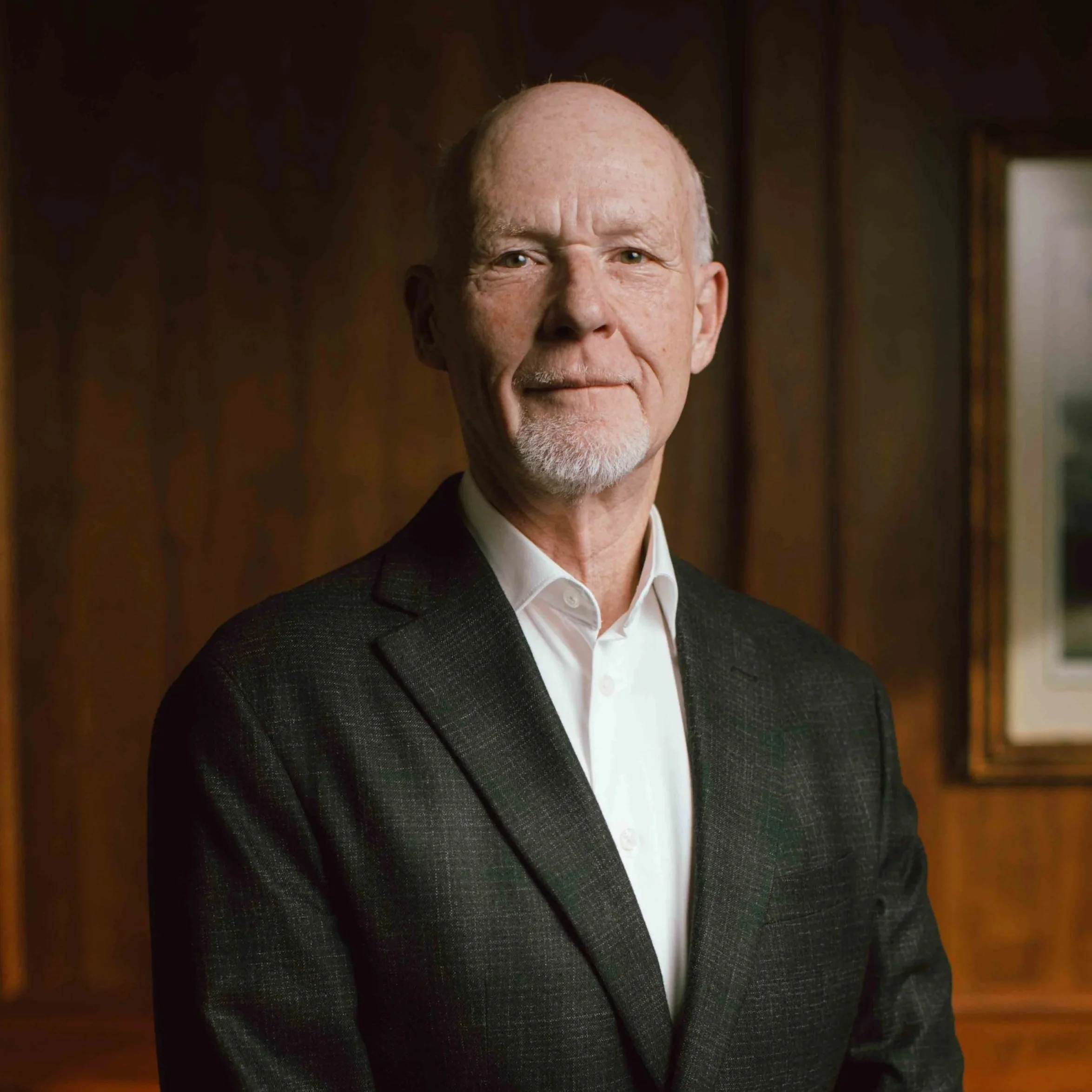 A close-up image of an older man with a bald head, facial hair, wearing a white shirt and a dark blazer, standing in front of a blurred background.