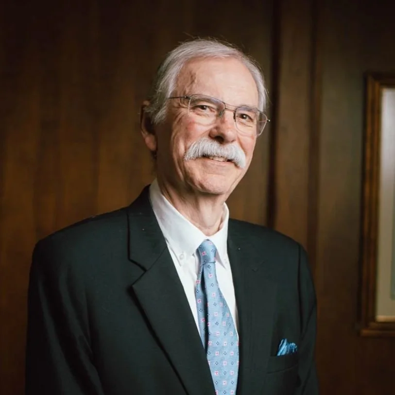 Portrait of an elderly man with gray hair, glasses, and a white mustache, wearing a black suit, white shirt, and a light blue tie with small patterns, standing indoors against a wooden wall background.