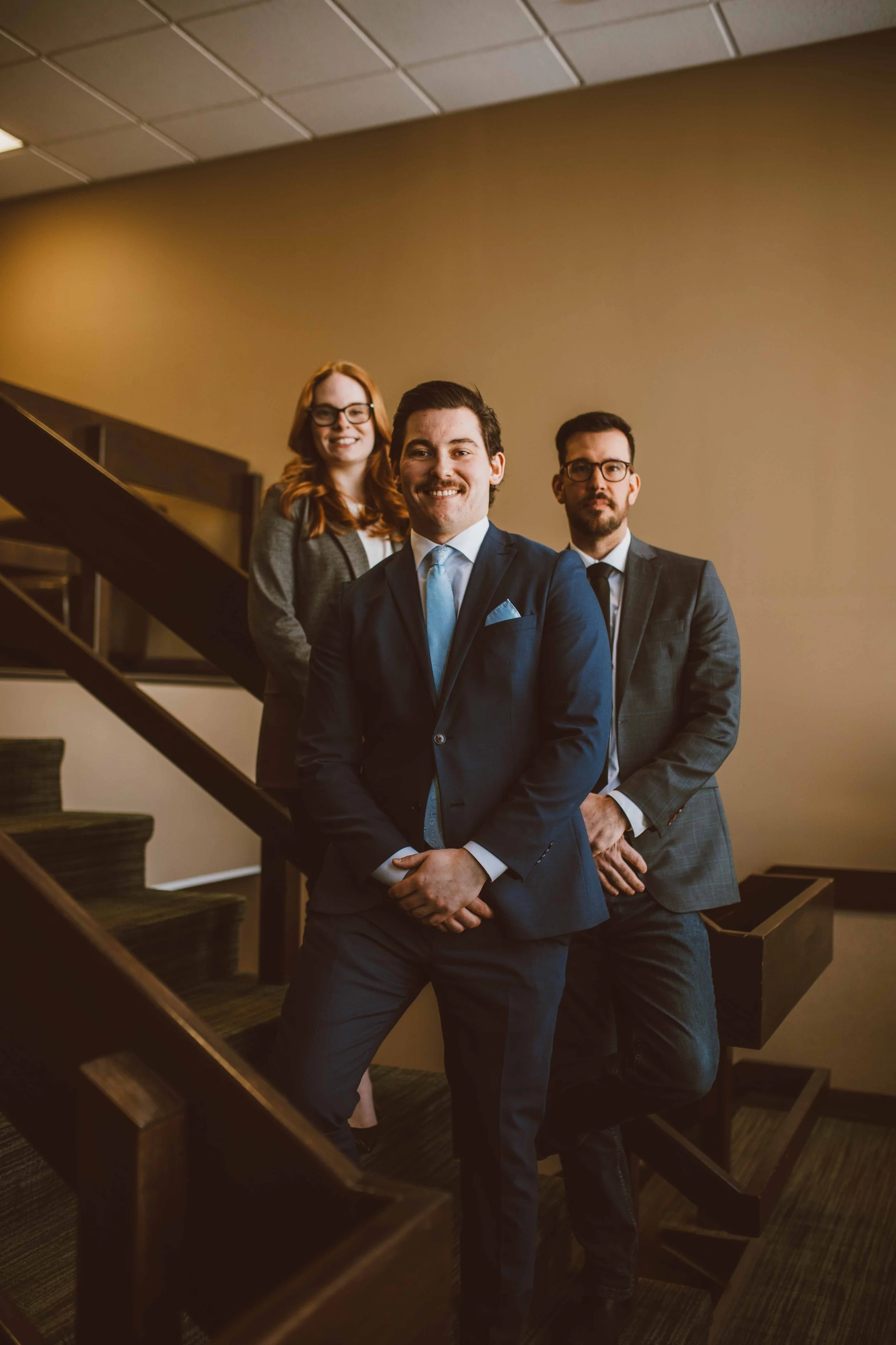 Three professionally dressed individuals, two men and one woman, stand on a staircase with a warm-colored wall in the background. The man in front is smiling and wearing a suit with a light blue tie. The woman and the other man are behind him, both wearing glasses and suits, with neutral expressions.