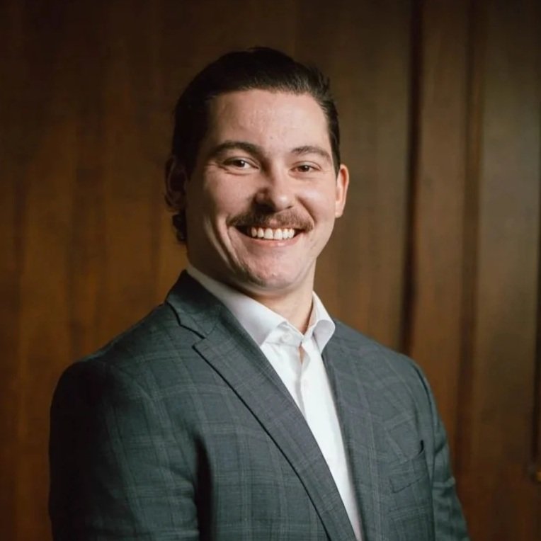 A young man with short dark hair, a mustache, wearing a suit jacket and white shirt, smiling against a wooden background.