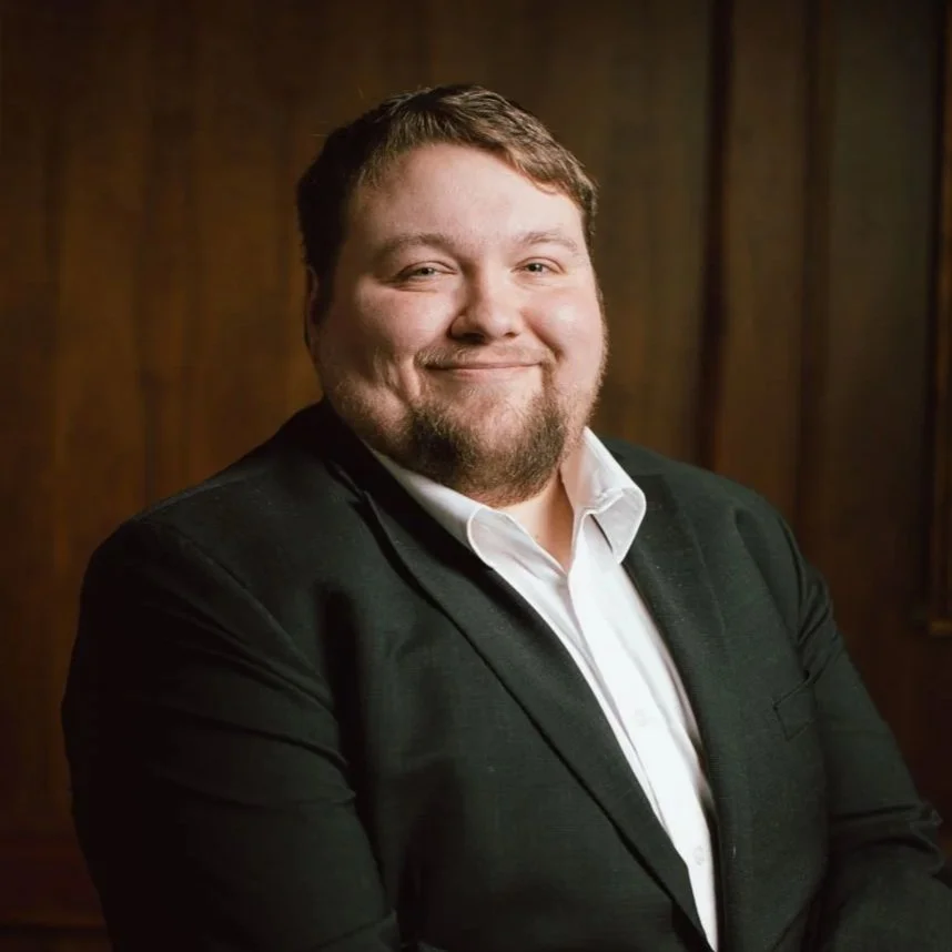 A man with light skin, short brown hair, and facial hair, wearing a black suit jacket and a white shirt, smiling and standing against a wooden background.