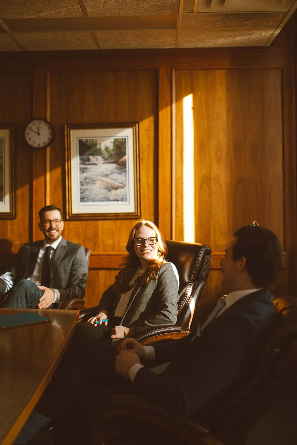 Three people, two men and one woman, are sitting and smiling in a wood-paneled conference room with framed artwork and a clock on the wall, sunlight streaming in.