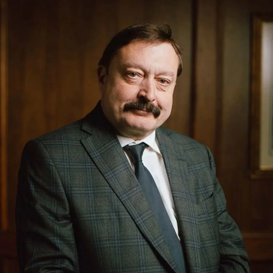 A man with a mustache and brown hair, wearing a dark plaid suit, white shirt, and dark tie, standing in a wood-paneled room.