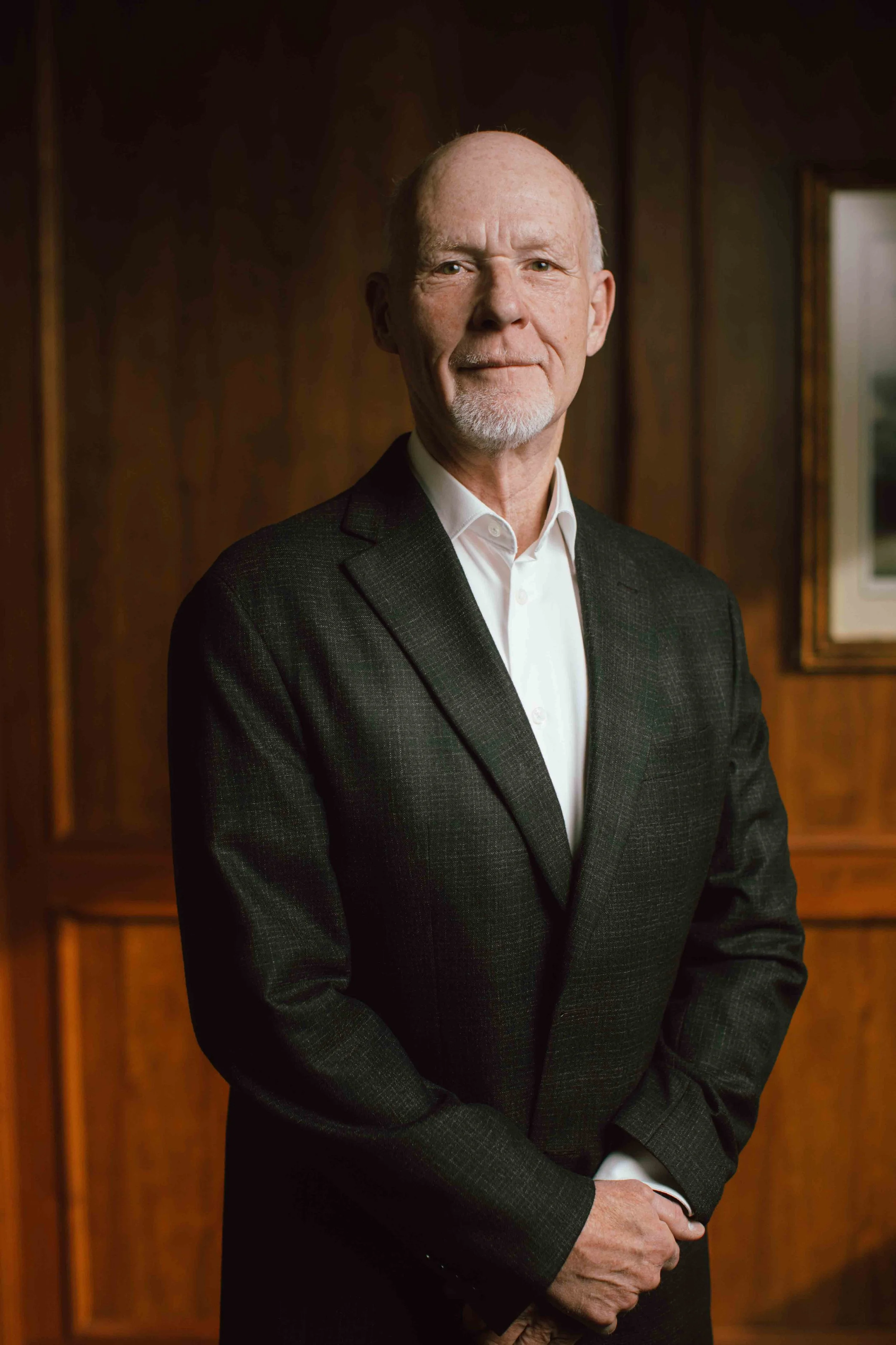 A portrait of an elderly man with a bald head, white beard, and mustache, dressed in a dark suit with a white shirt, standing in front of a wood-paneled wall with a framed painting visible on the right.