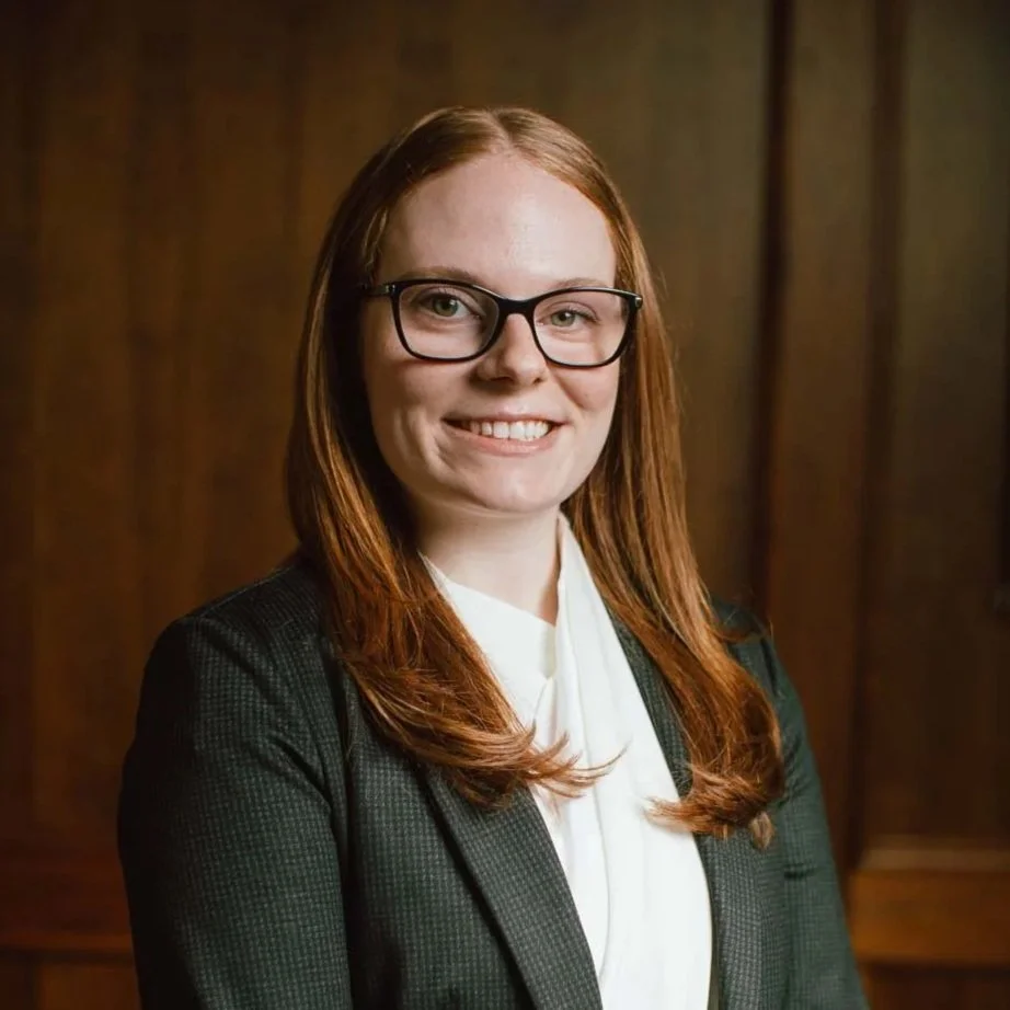A woman with long red hair and glasses, wearing a dark blazer and white blouse, smiling in front of a wooden background.