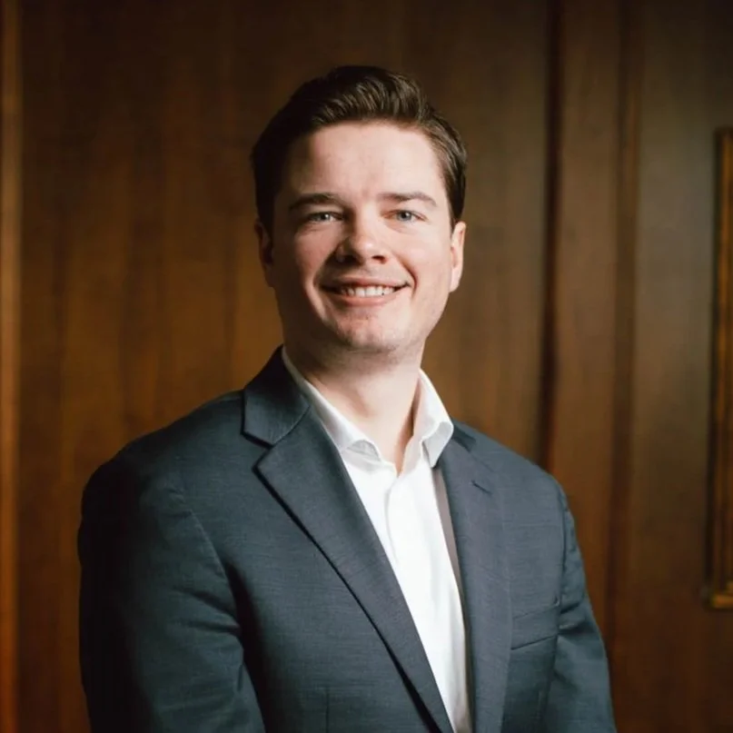 A young man with dark hair, wearing a suit and white shirt, smiling and standing in front of a wooden wall.