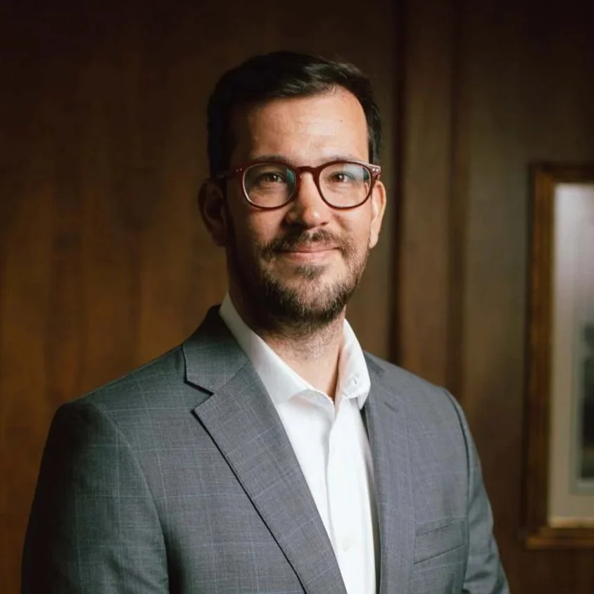 A man with glasses and a beard wearing a gray suit and white shirt, smiling in an indoor setting with wooden panel walls.