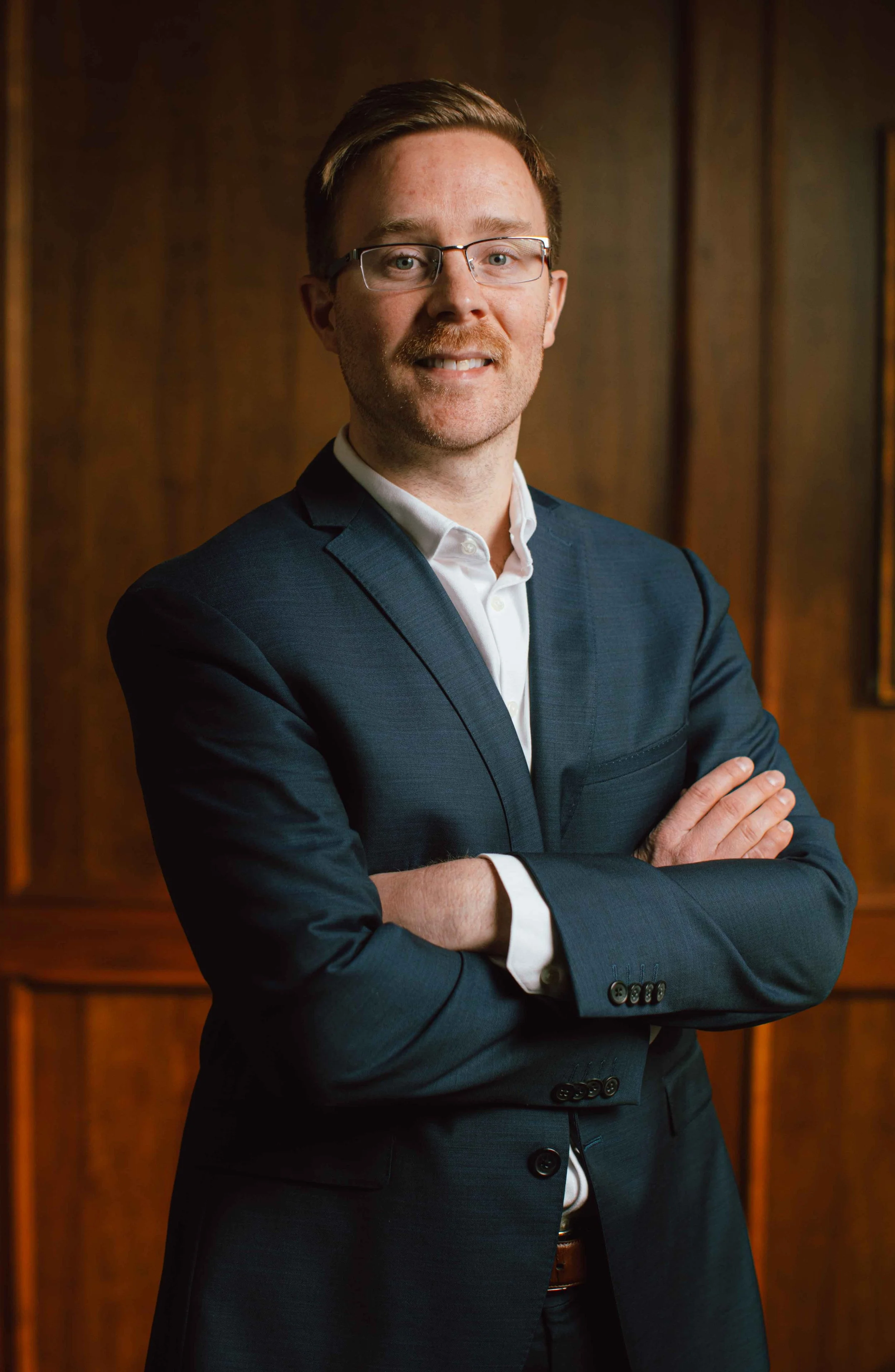 Man in a navy suit and glasses standing with arms crossed in a wood-paneled room.