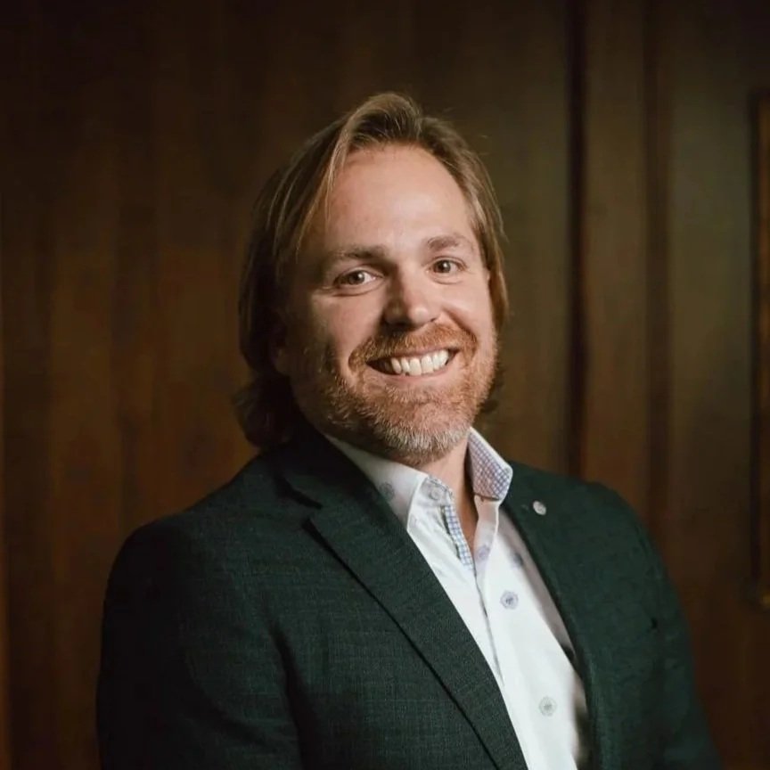 A man with shoulder-length blond hair and a beard, smiling, wearing a dark blazer over a white collared shirt, standing against a wooden-paneled background.