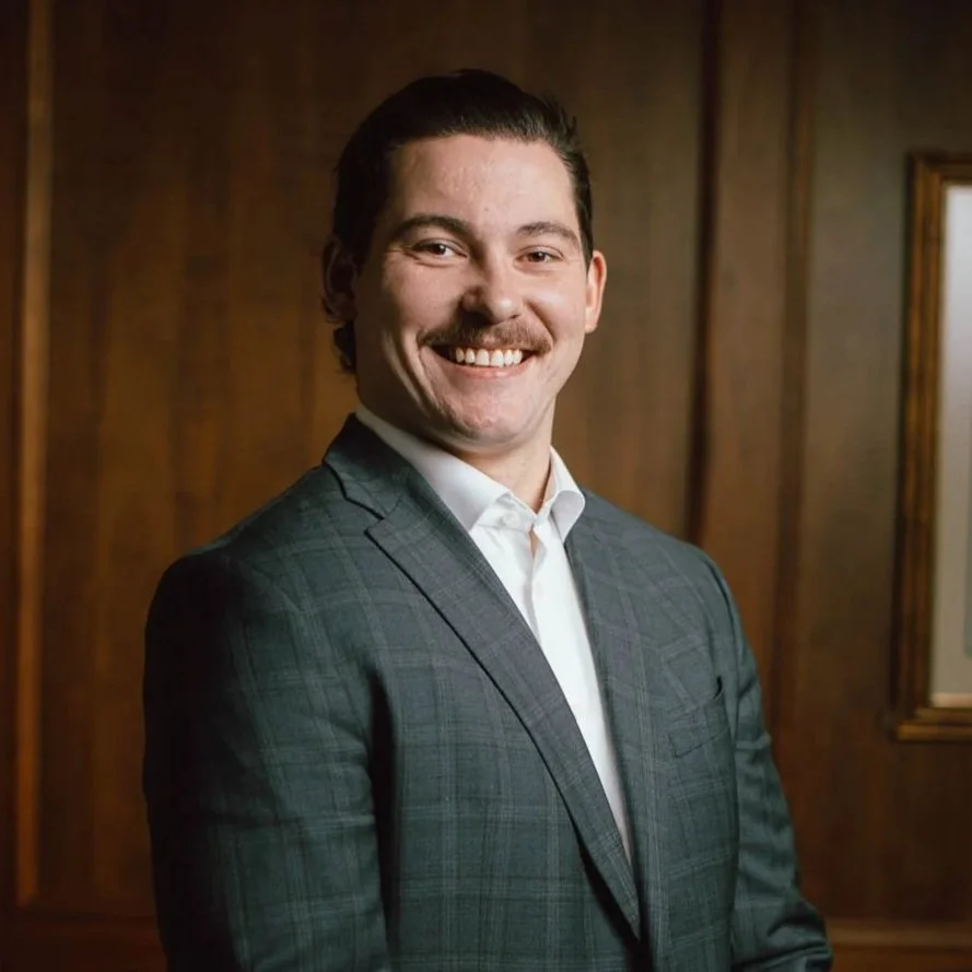 A man with dark hair, mustache, and light skin, wearing a checked suit and white shirt, standing in front of wooden paneling, smiling at the camera.