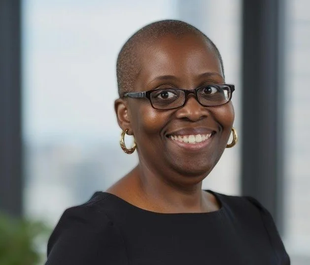 Close-up of an African American woman with a shaved head, wearing glasses, gold hoop earrings, and a black top, smiling in an office setting.