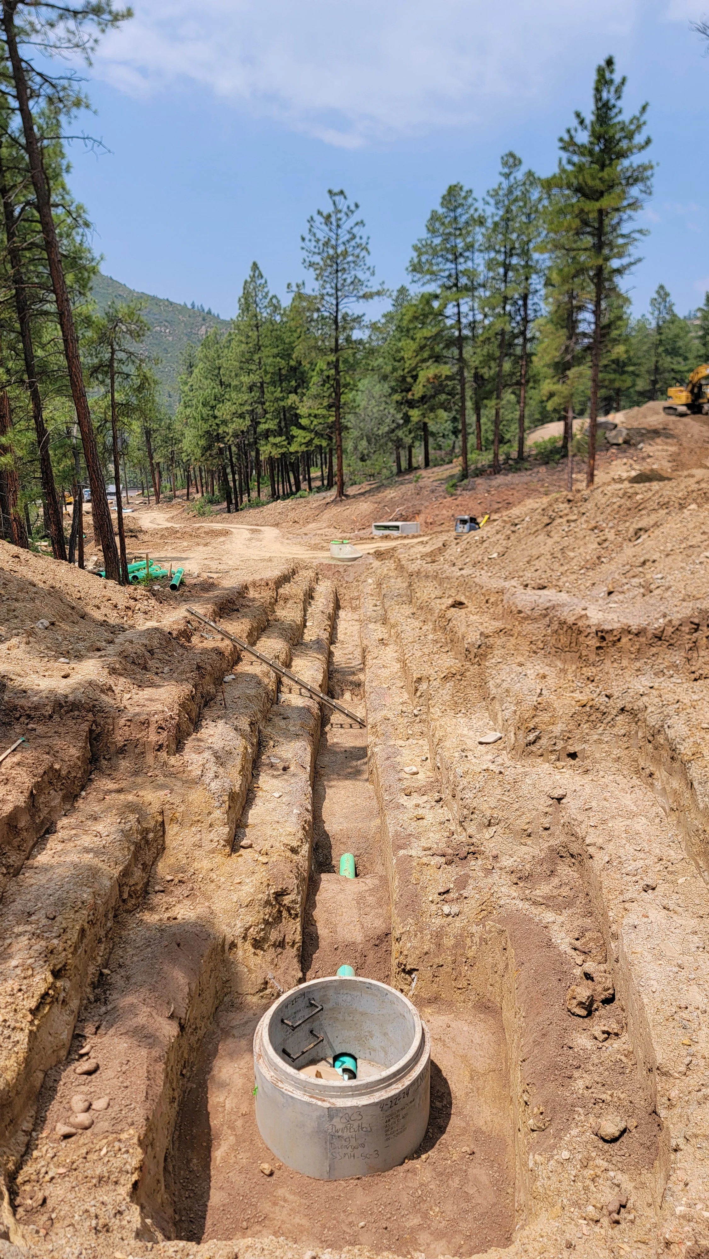 Construction site with trenches and pipes in a forested area, with trees and a blue sky in the background.