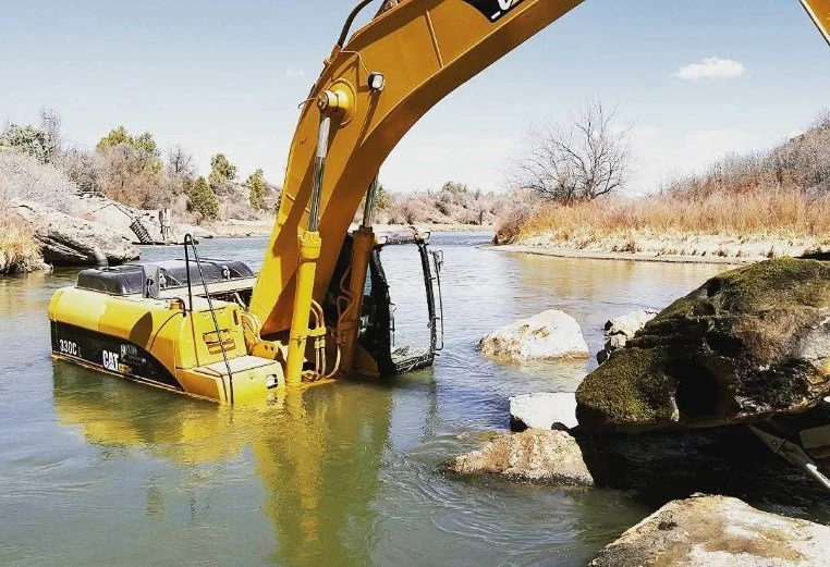 A yellow Caterpillar excavator partially submerged in a river, with rocks and a natural landscape in the background.