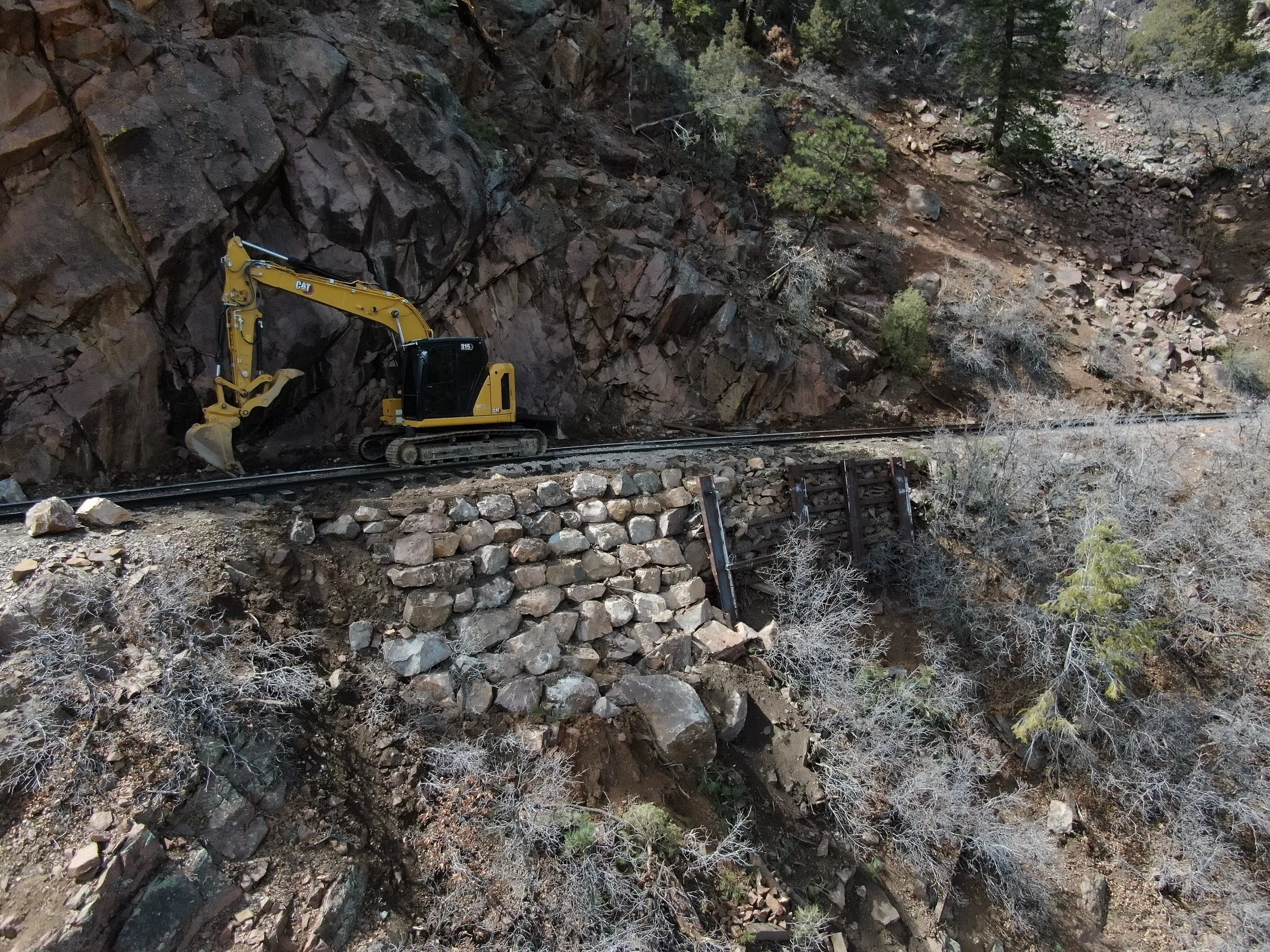 A small yellow Caterpillar excavator working on a mountainside next to railroad tracks, with a stone retaining wall and sparse vegetation.