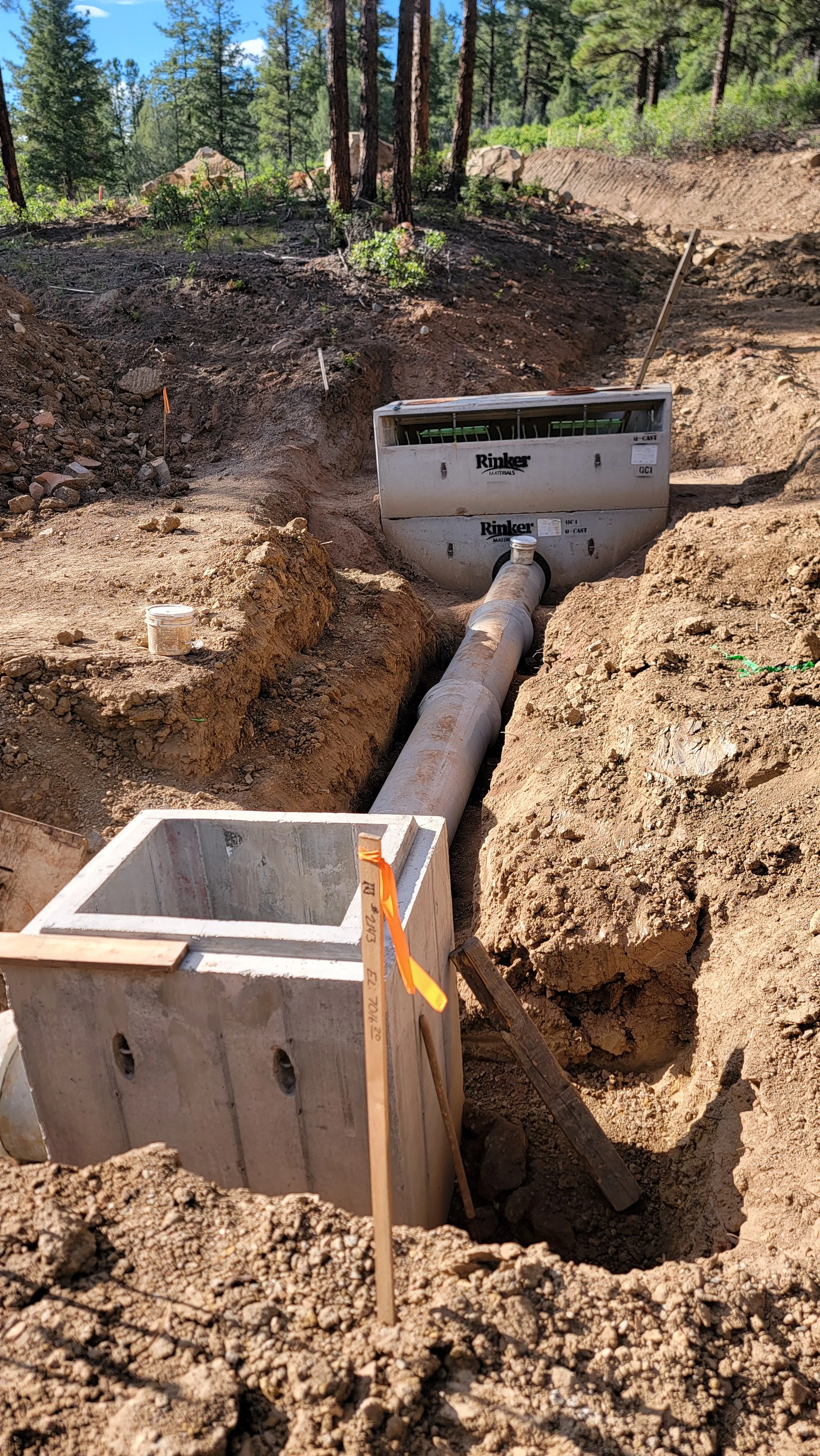 Construction site with a large pipe extending into a trench surrounded by dirt and trees in the background, with equipment and markers visible.