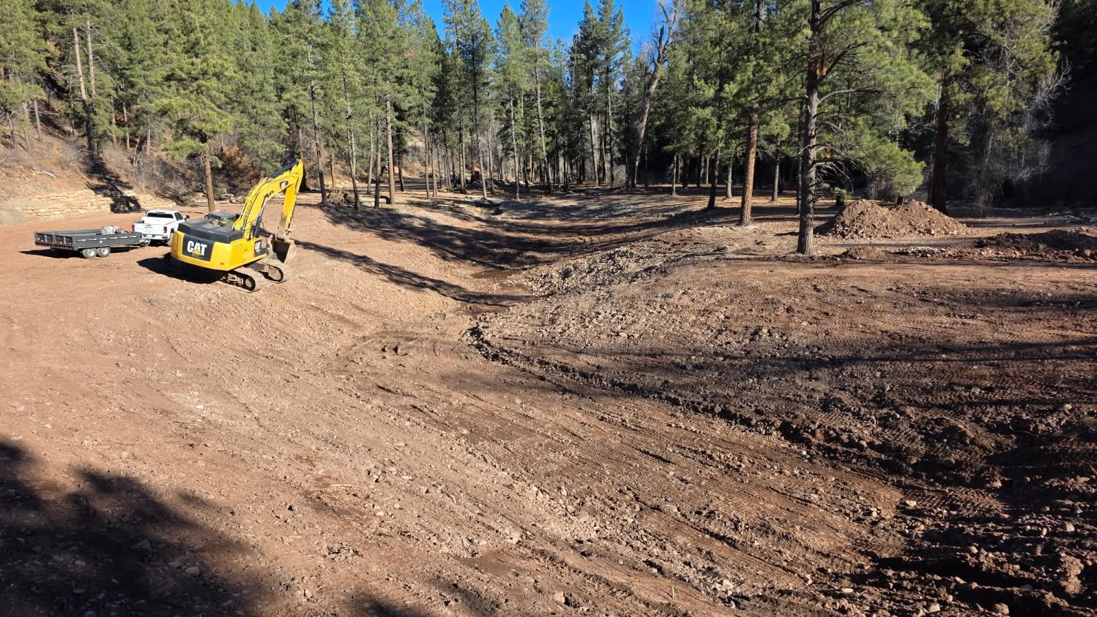 A construction site in a wooded area with a yellow CAT excavator, a white pickup truck, and a trailer, with a cleared dirt area and tall trees in the background.