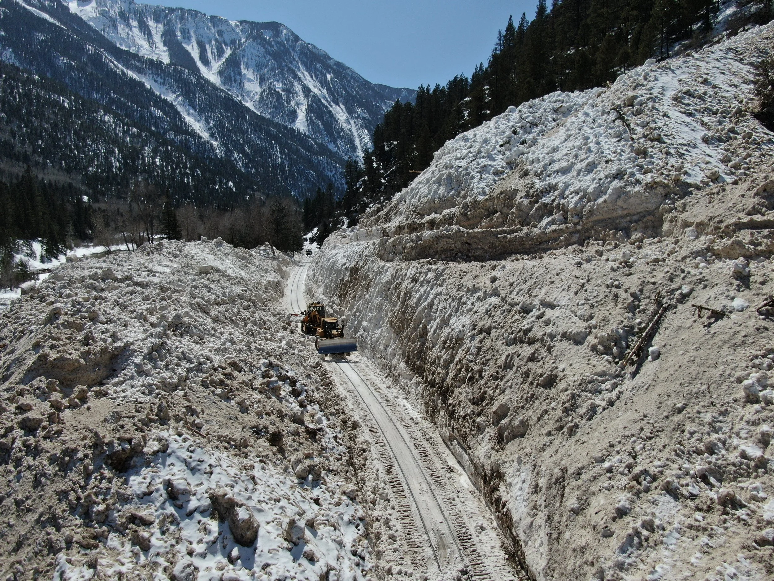 Construction workers are clearing snow from railway tracks in a mountainous area with snow-covered slopes and pine trees.