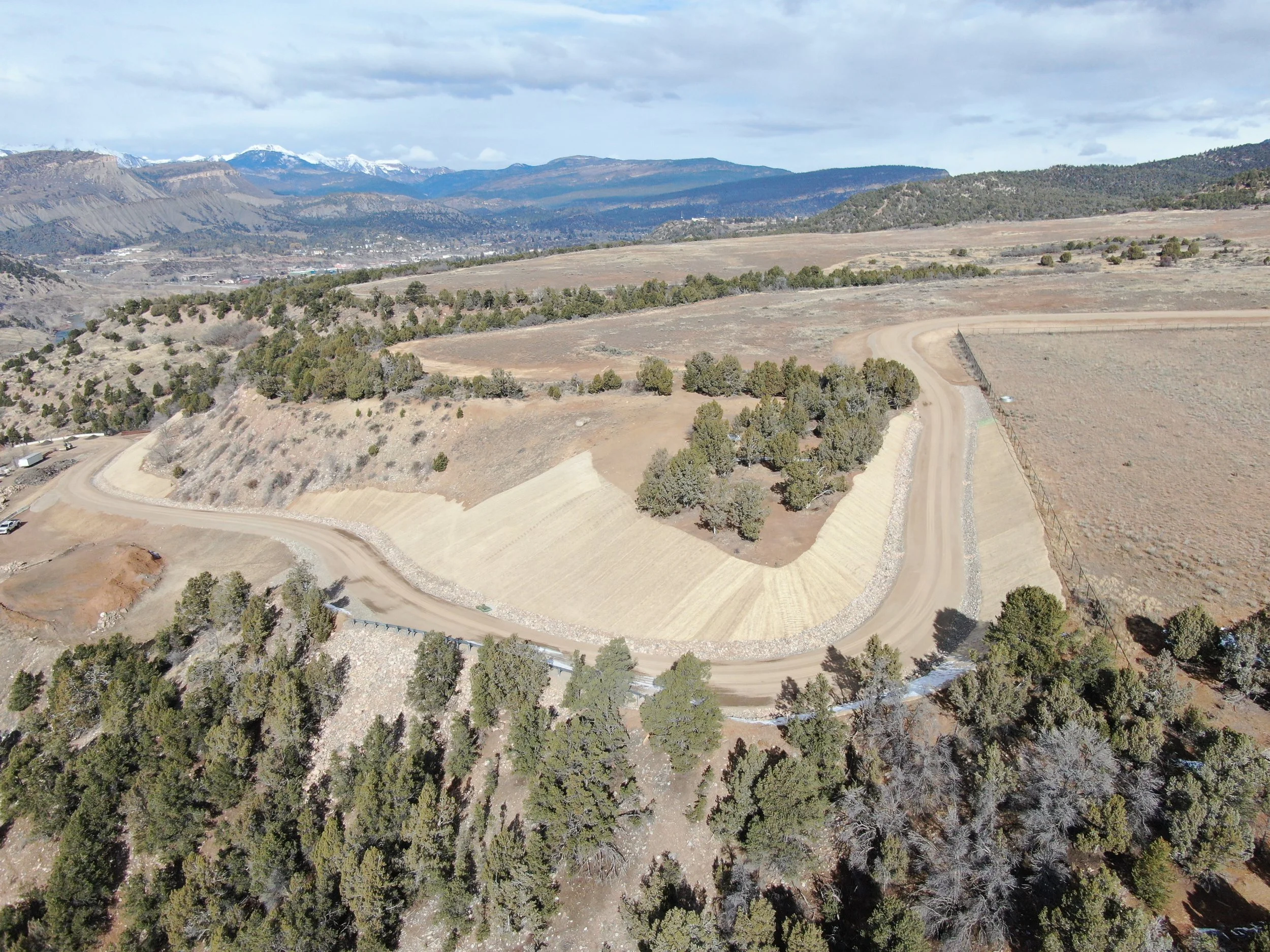 Aerial view of a winding dirt road through a mountainous landscape with trees, hills, and snow-capped mountains in the background.