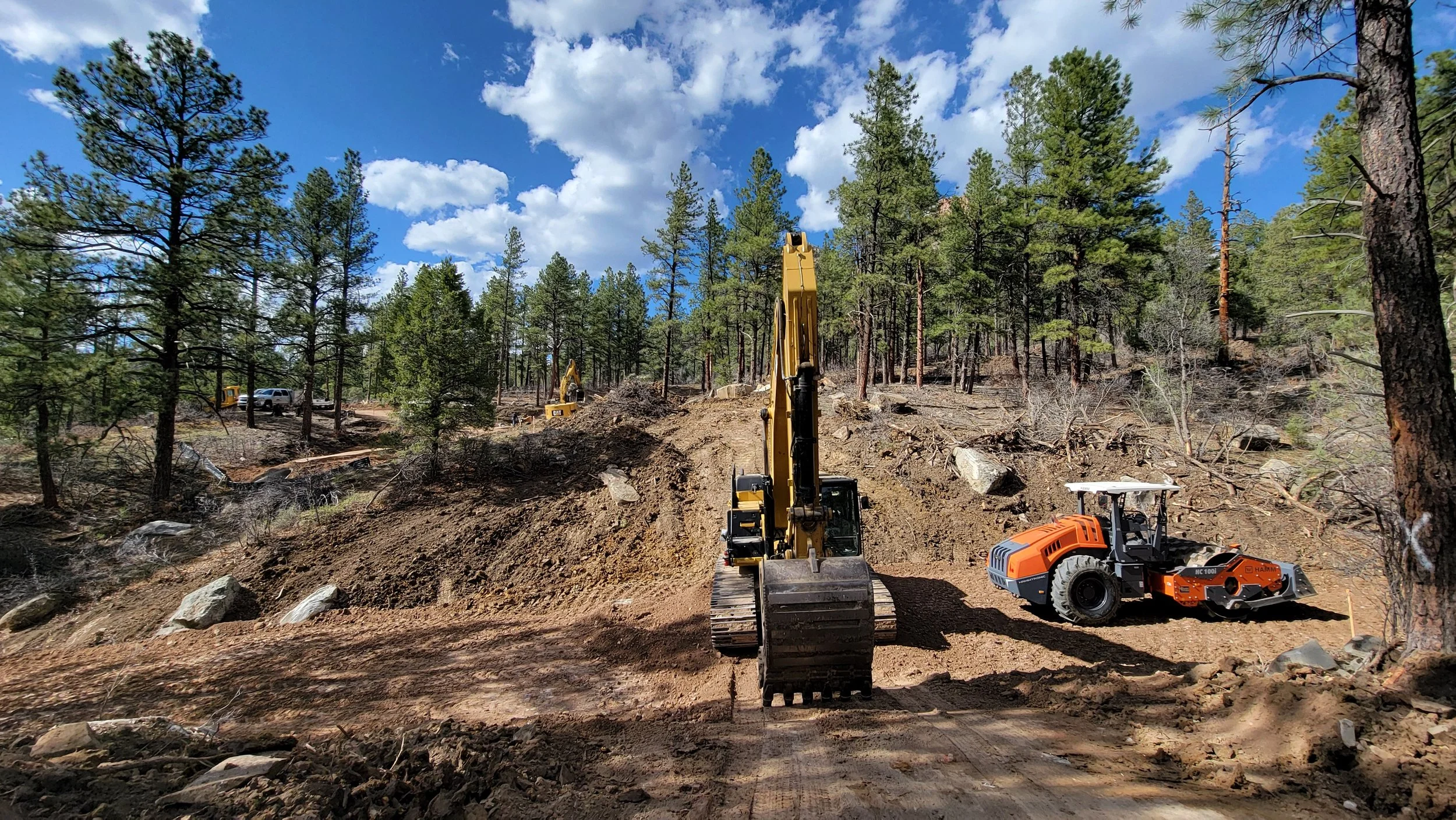 Construction site in a forest with excavators and a roller compactor working on grading and clearing the land, under a partly cloudy sky.