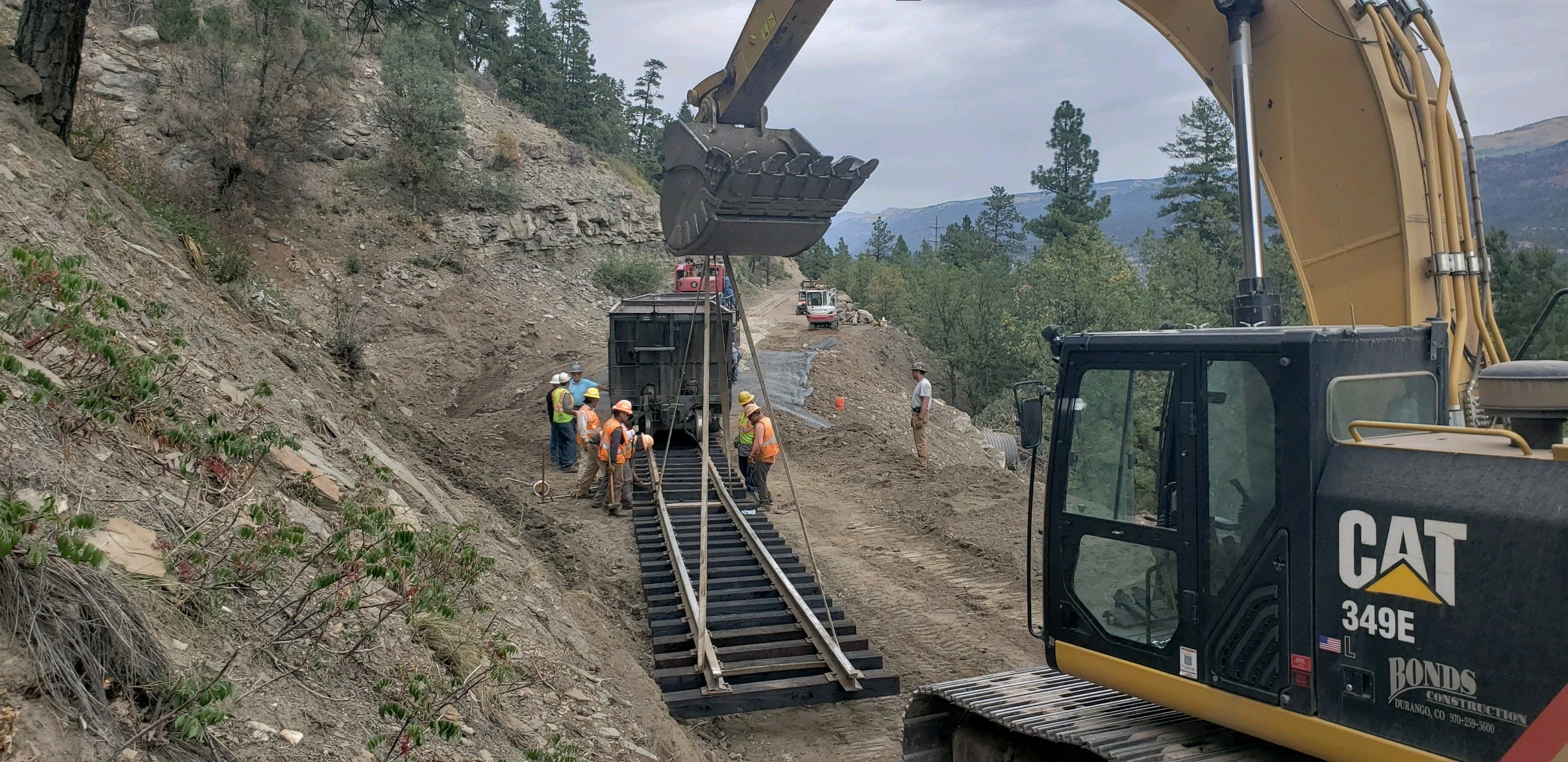Construction workers in hard hats and safety vests working on a mountain hillside, using a large excavator to lay railroad tracks.