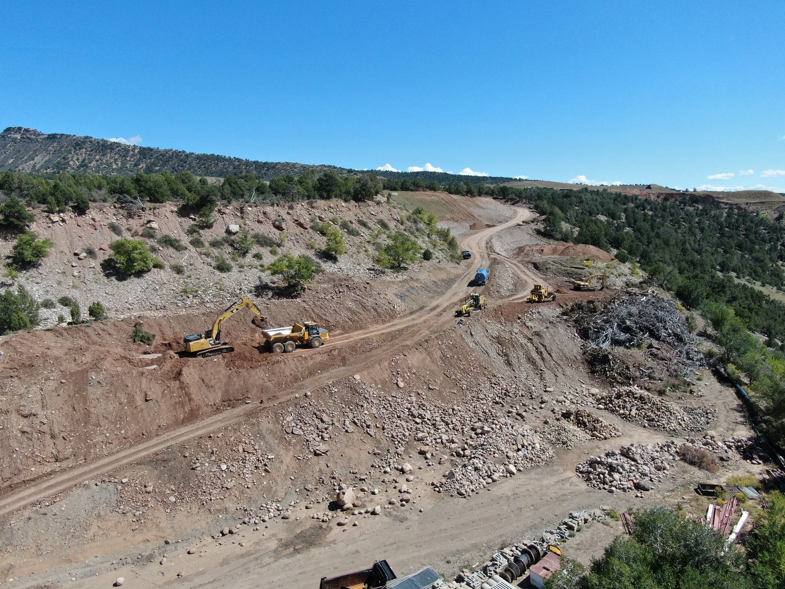 Construction site in a hilly, semi-arid landscape with excavators, trucks, and machinery working on the hillside under a clear blue sky.