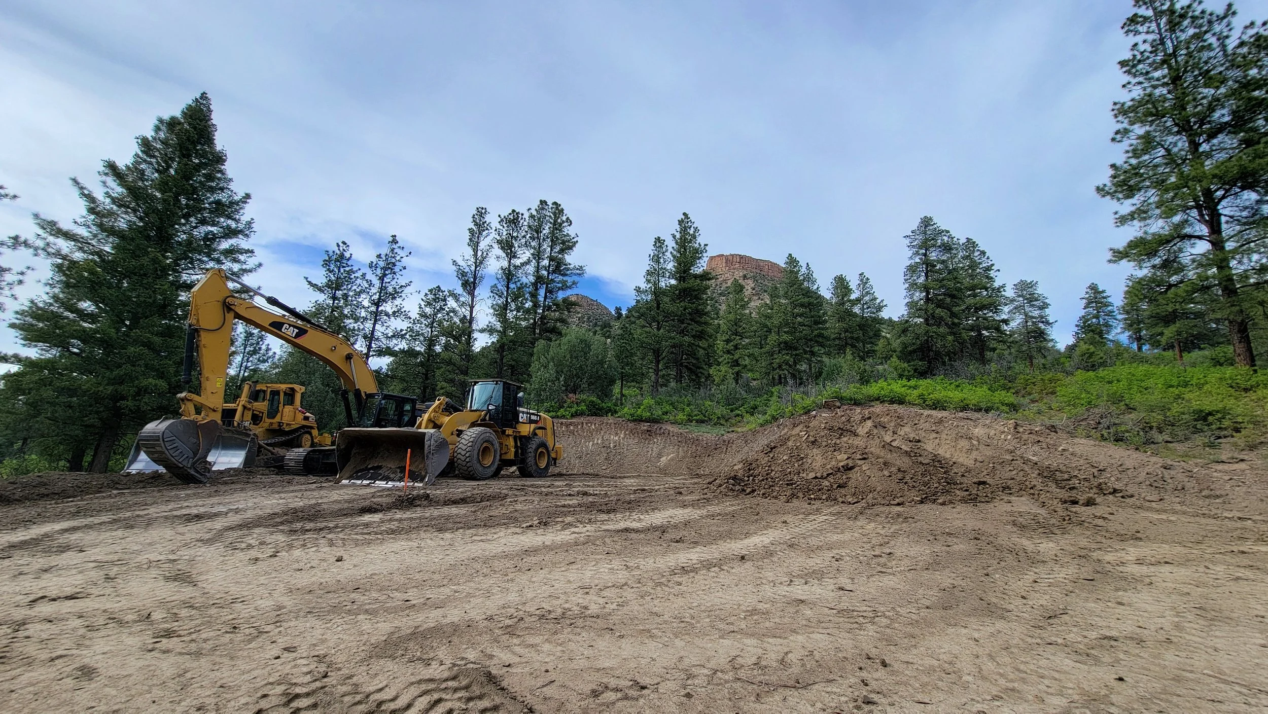 Construction site with yellow bulldozers working on dirt terrain surrounded by trees and mountains in the background.