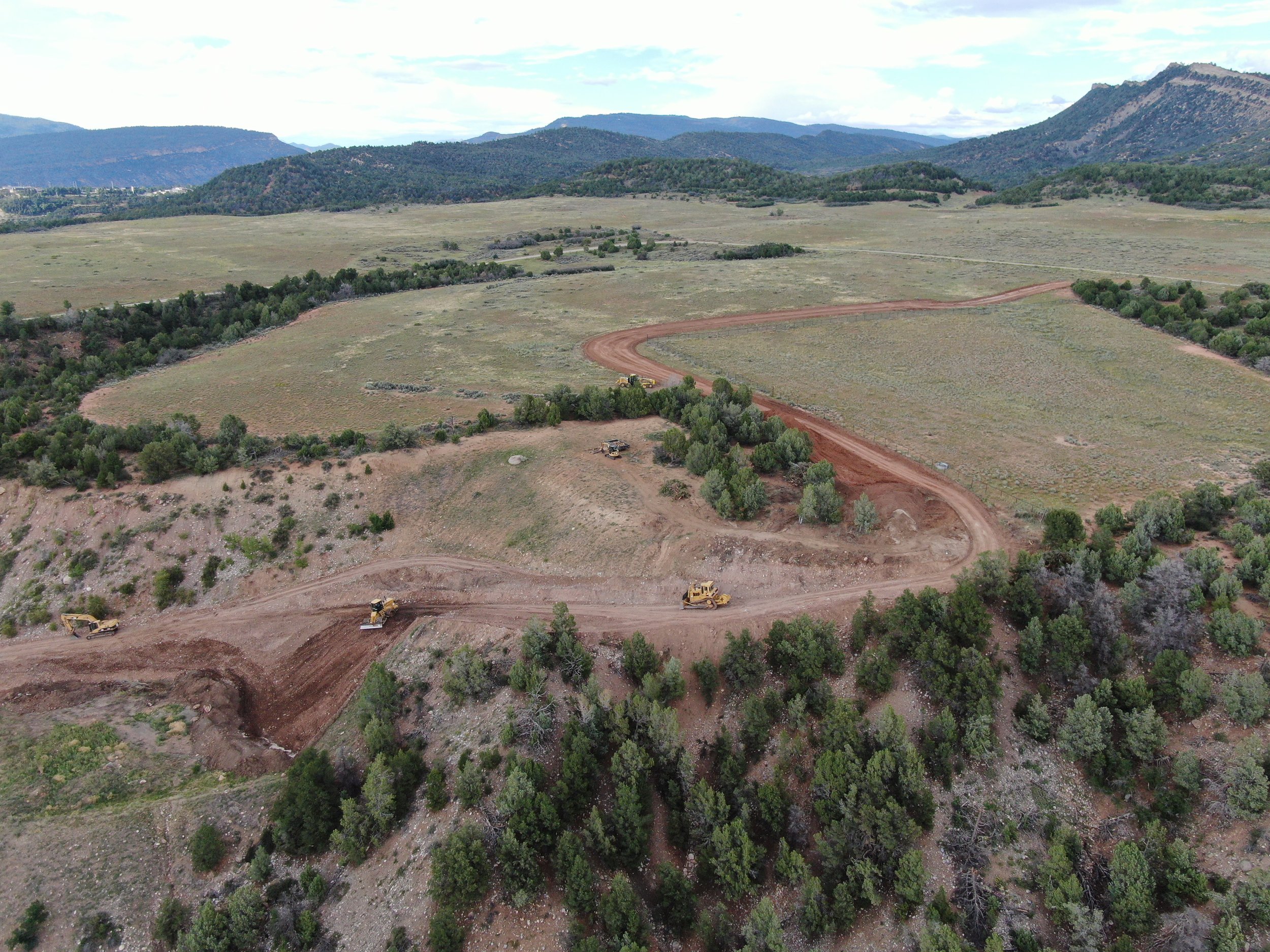 Aerial view of a construction site in a hilly, rural landscape with dirt roads and several construction vehicles working on grading and clearing land, surrounded by green trees and expansive fields with mountains in the background.