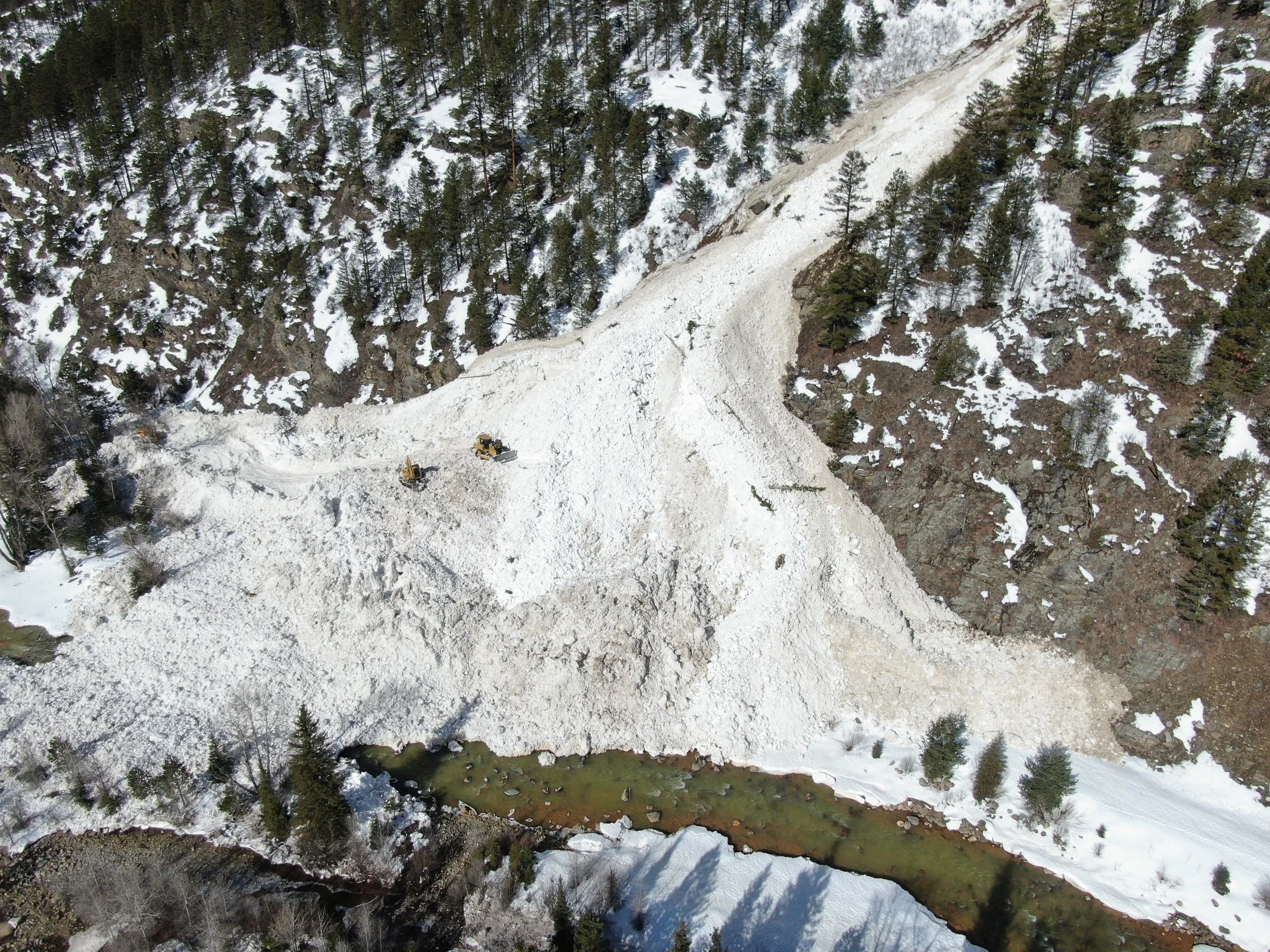 Snow removal equipment clearing snow on a mountain road with trees, rocks, and a small creek below.