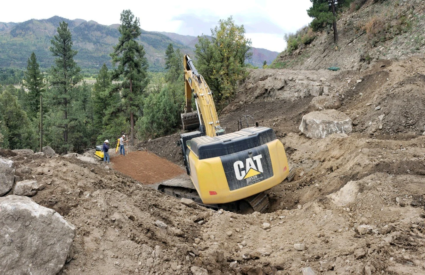 Construction site on a hillside with a yellow CAT excavator and workers laying down a dirt path surrounded by trees and mountains in the background.