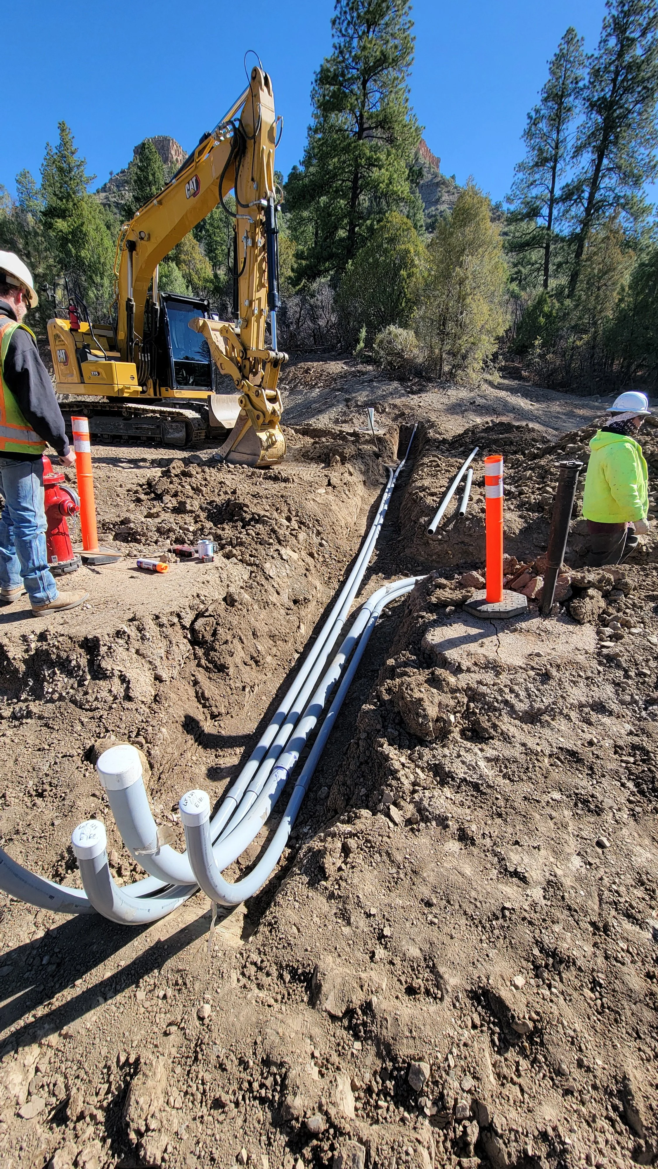 Construction site with a yellow excavator, white electrical pipes laid in the dirt, and workers in safety vests and helmets working on underground utility installation amid trees and mountains in the background.