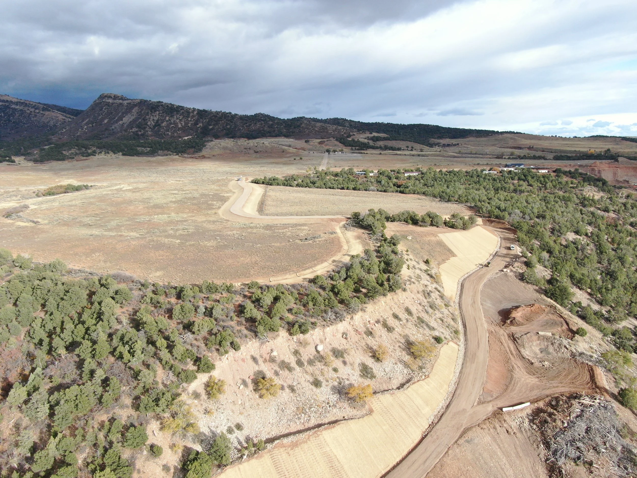 Aerial view of a hilly landscape with dirt roads winding through patches of green shrubbery and trees, and a mountain range in the background under a cloudy sky.